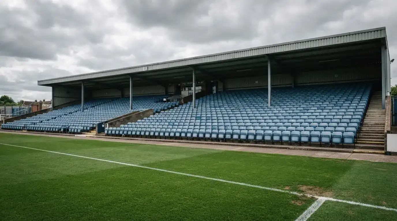 Rzędy pustych plastikowych krzesełek małego stadionu widziane z poziomu murawy pod pochmurnym niebem.