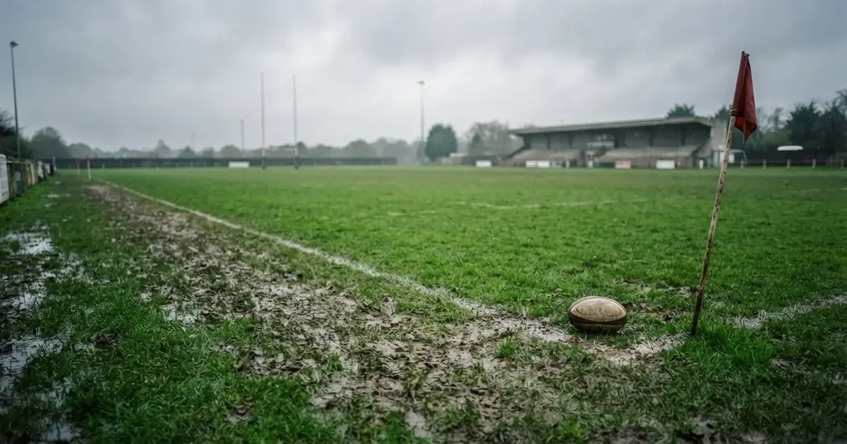 Boisko do rugby ze słupami bramki w oddali pod pochmurnym niebem, mokra trawa i ślady butów.