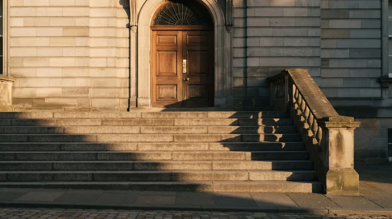 Granitowe schody prowadzące do wysokich drewnianych drzwi budynku w porannym świetle, pusta balustrada.