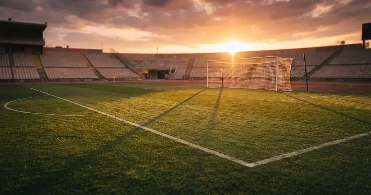 Murawa stadionu o zachodzie słońca z długimi cieniami słupków bramki, trybuny w ciepłym świetle.
