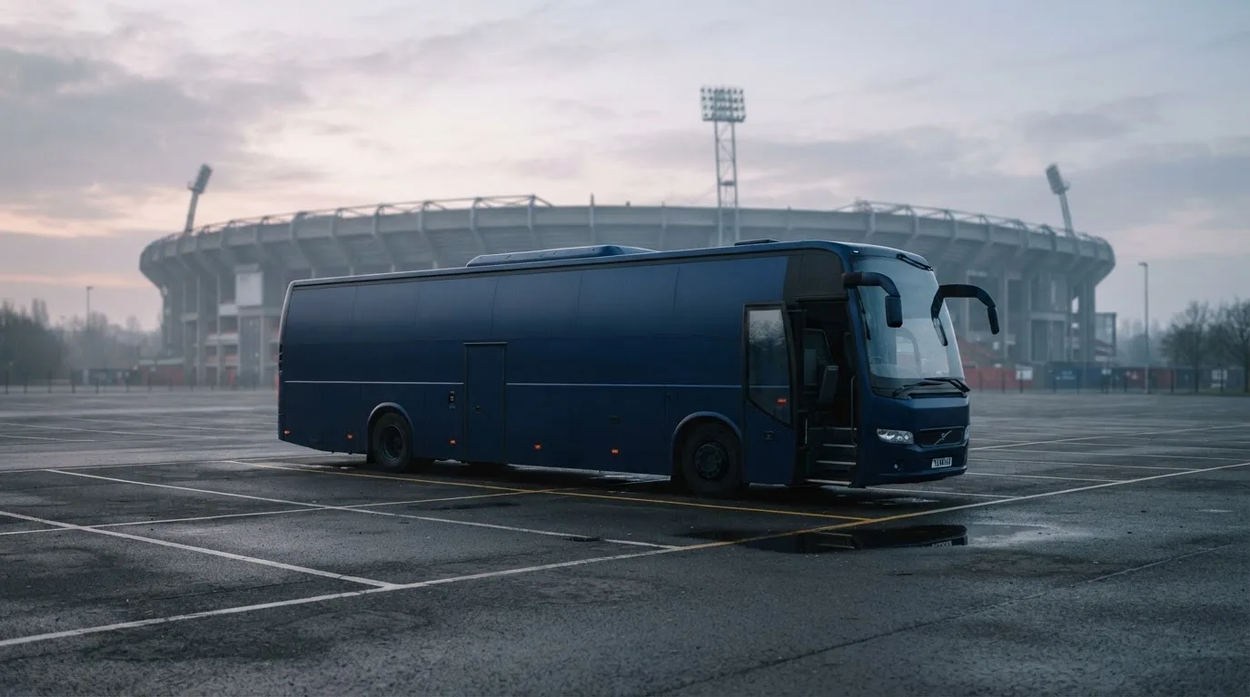 Autokar stoi na parkingu przed stadionem o świcie, rozmyte kontury trybun w tle, pusty plac i mokry asfalt.